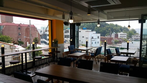 An indoor dining area with tables and chairs, large windows offering a city view, and hanging pendant lights. - The View Hostel (Tongyeong)