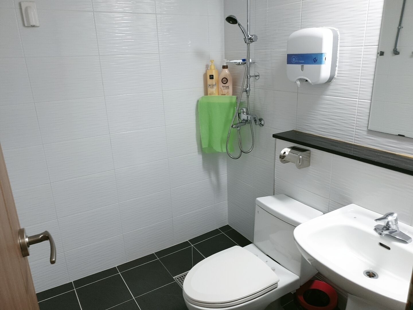 A bathroom with a white toilet, a sink, and a shower. There are toiletries on the wall and a green towel hanging.