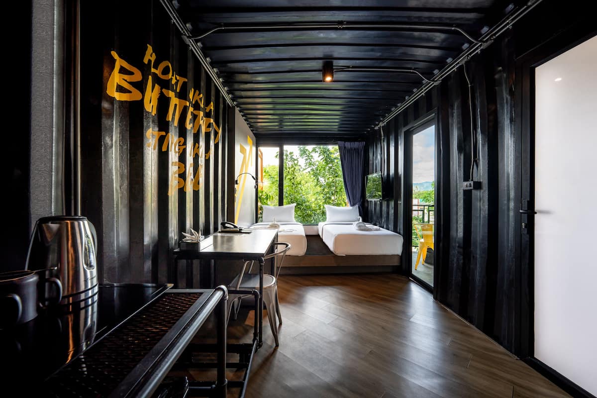 A modern room with a wooden floor, a bed, a table with a kettle, and a window with a view of greenery.