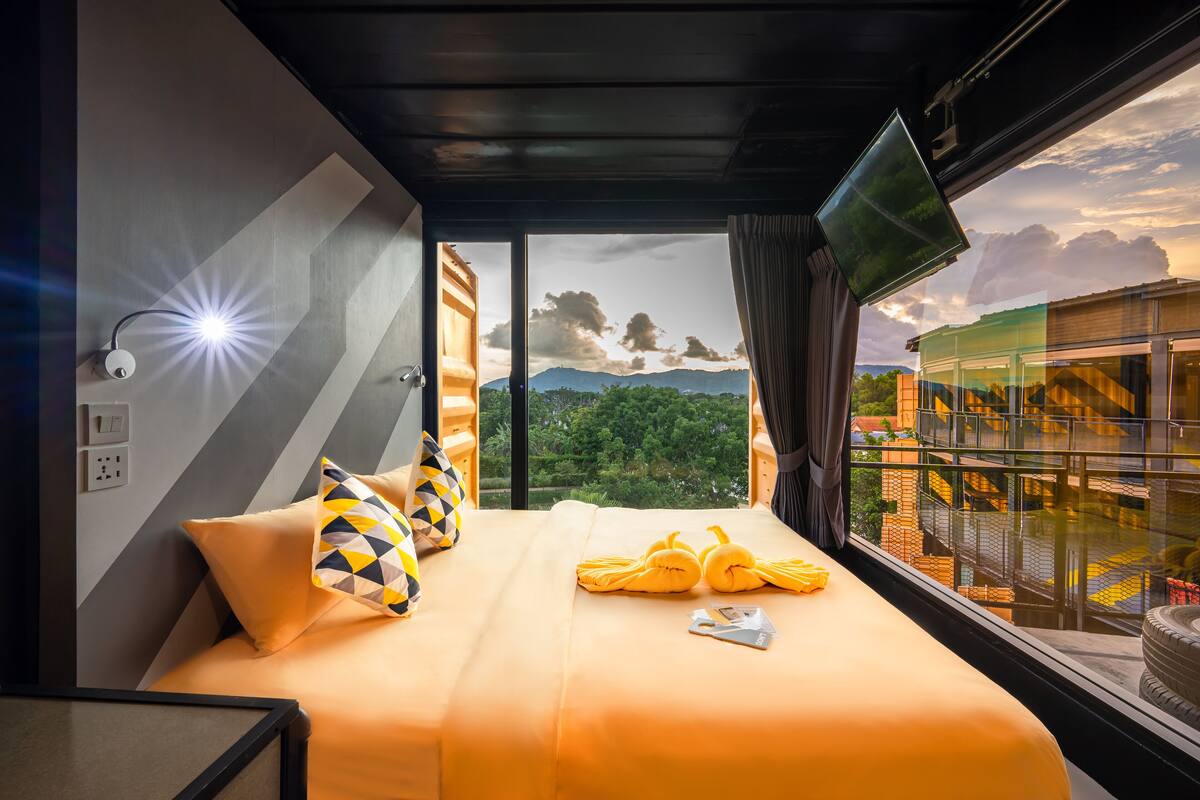 Una habitación de hotel con cama, almohadas, televisión y vistas a un edificio y montañas.