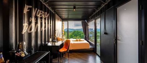 A modern hotel room with a large bed, a desk with a chair, a coffee machine, and a view of the outdoors.