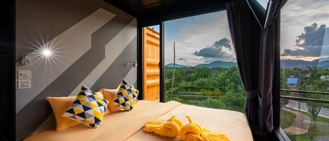A hotel room with a large bed, two pillows, a towel, and a window with a view of greenery and mountains.