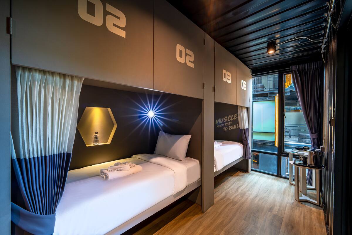 Una habitación de hotel moderna con dos camas, una mesita, una lámpara y vistas a la ciudad a través de amplios ventanales.