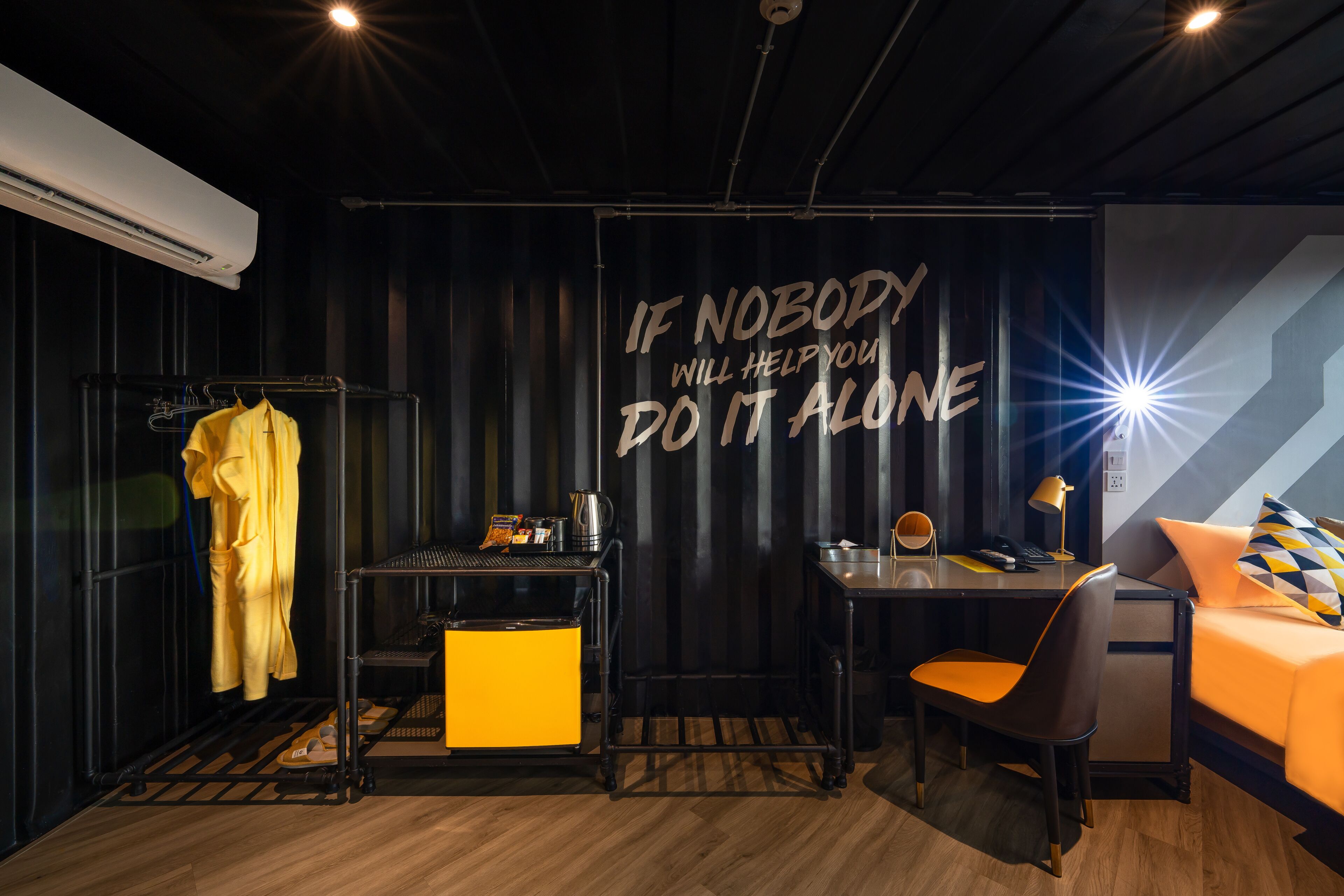 A modern room with a black and yellow color scheme, a motivational quote on the wall, a desk with a chair, a bed, and a hanging rack with clothes.