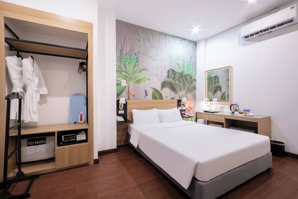 A hotel room with a bed, a wardrobe with hanging robes, a desk with a lamp, and a wall-mounted air conditioner.