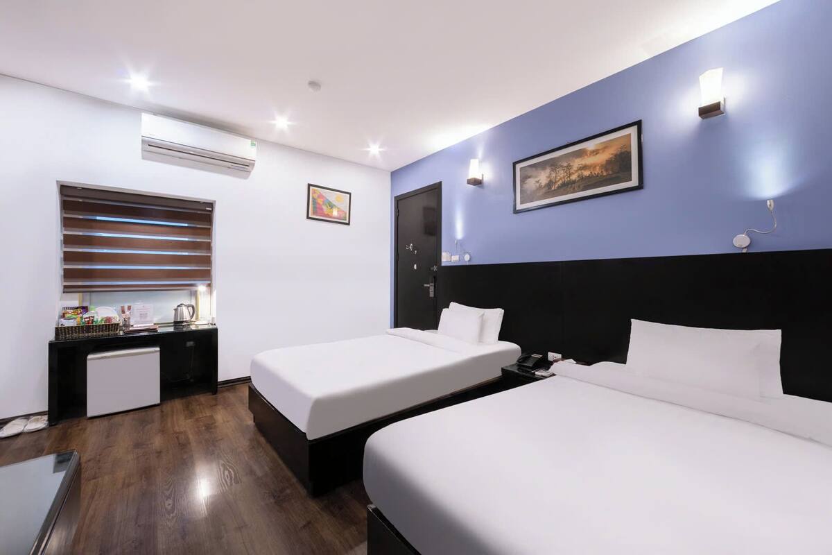 A hotel room with two beds, a small kitchenette, and a window with blinds.