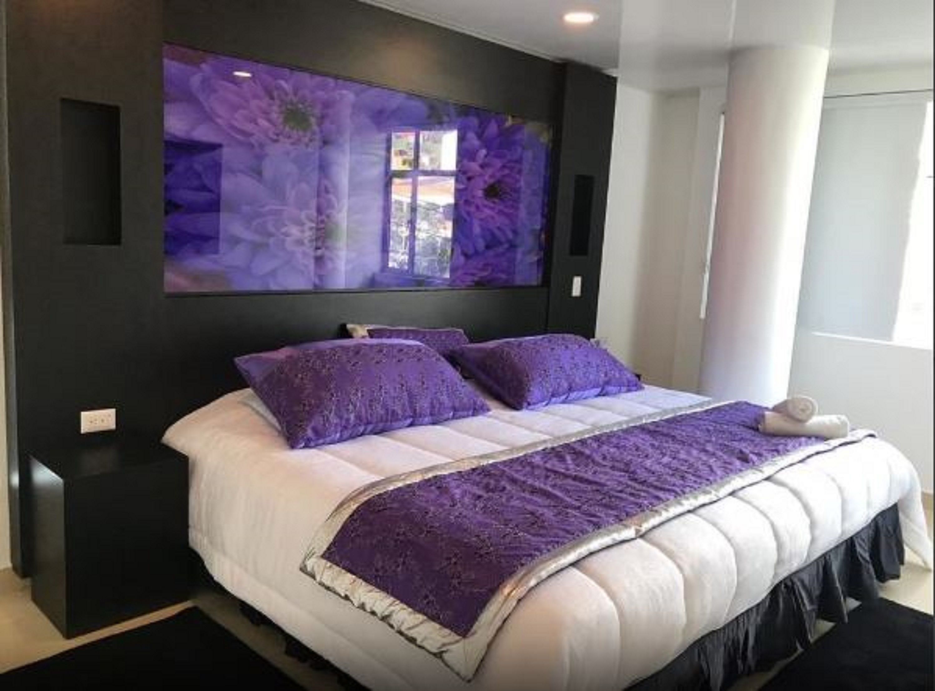 Un dormitorio moderno con una cama grande, ropa de cama morada y una obra de arte floral de gran tamaño en la pared.