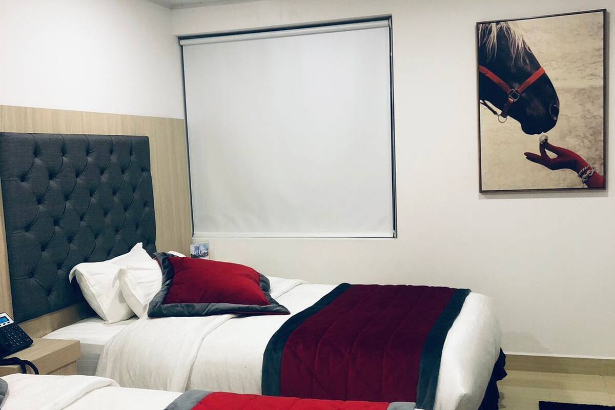 Habitación de hotel con dos camas, cada una con cubrecama acolchado rojo y blanco, cabecero gris y un ventanal grande con persiana blanca.