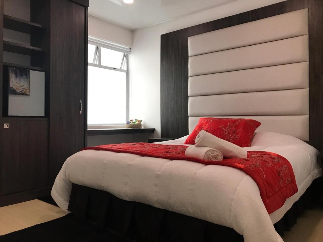 Una cama bien hecha con una colcha y almohada rojas, un armario de madera y una ventana con luz natural.