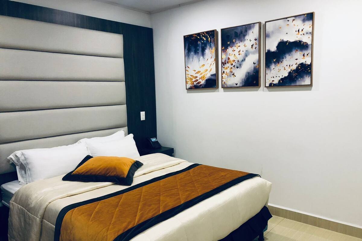 Una habitación de hotel moderna con una cama grande, un cabecero con líneas horizontales, una mesita de noche con un teléfono y tres cuadros abstractos en la pared.