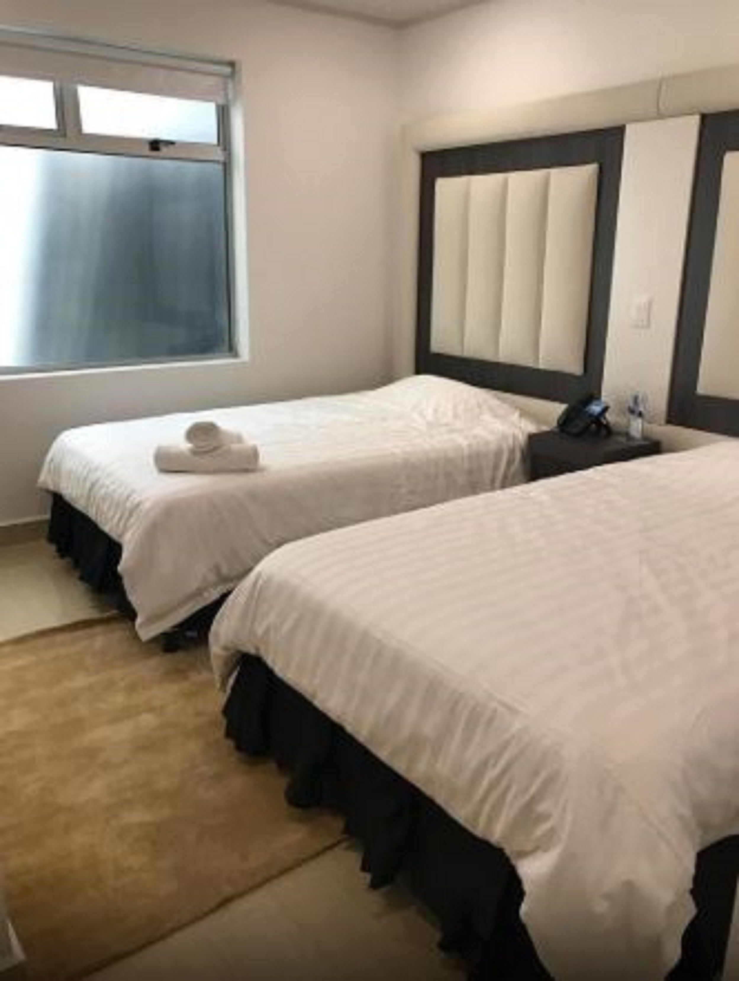 A hotel room with two single beds, a large window, and a headboard with a light-colored panel.