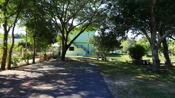 A paved pathway in a park with trees, a bench, and a building with a staircase in the background.