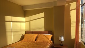 Una cama bien hecha con una colcha estampada y almohadas, una mesita de noche con una lámpara y una ventana con cortinas.