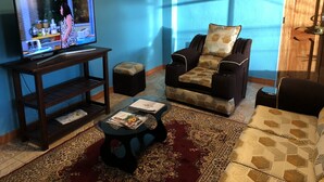 Una sala de estar con televisor sobre un soporte de madera, un sillón estampado, un pequeño reposapiés y un tapete con diseño.