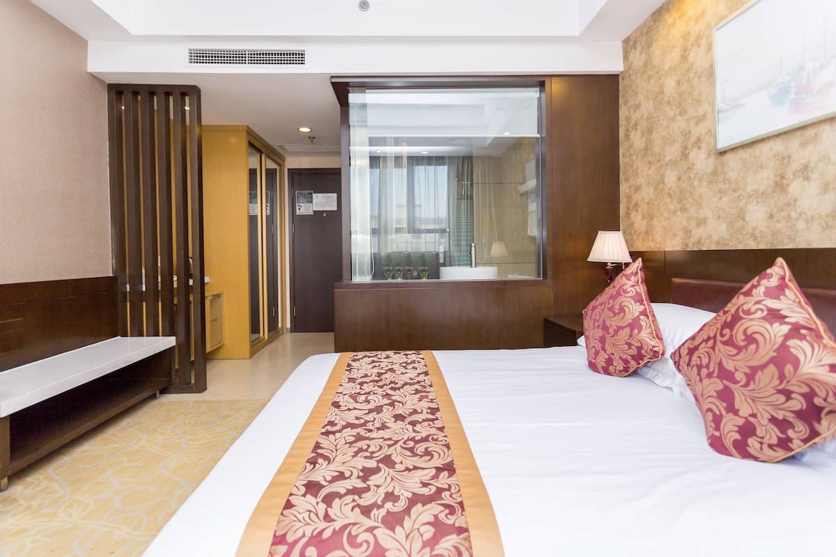 Une chambre d’hôtel avec un grand lit, une table de chevet, un banc, une fenêtre avec des rideaux et un téléviseur fixé au mur.