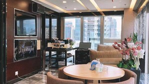 Um saguão moderno de hotel com uma mesa redonda, assentos confortáveis, uma televisão grande e vista da cidade através de amplas janelas.