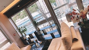 Um interior moderno com sofá e mesa, janelas amplas com vista para prédios e plantas decorativas.