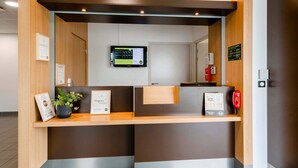 A reception desk with a flat-screen monitor, framed certificates, a potted plant, and a red telephone. - B&B Hotel Nantes Savenay (Savenay)