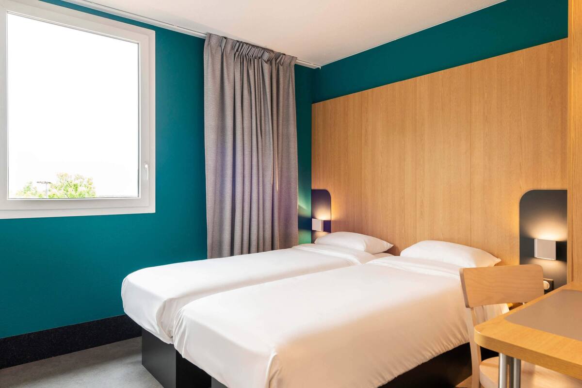 Hotellihuone, jossa on kaksi sänkyä, ikkuna, jossa on verhot, puinen pääty ja pöytä tuolilla.