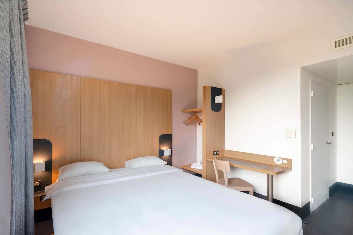 Hotellihuone, jossa on sänky, yöpöytä, lamppu, peili ja työpöytä tuolilla.