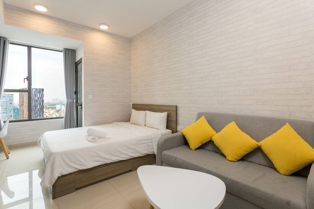 A modern hotel room with a bed, a gray sofa with yellow pillows, a white coffee table, and a large window offering a city view.