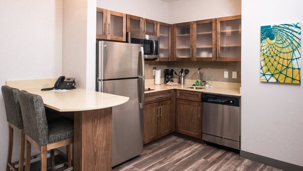 A compact kitchen with wooden cabinets, stainless steel appliances, a bar stool, and a wall-mounted artwork.