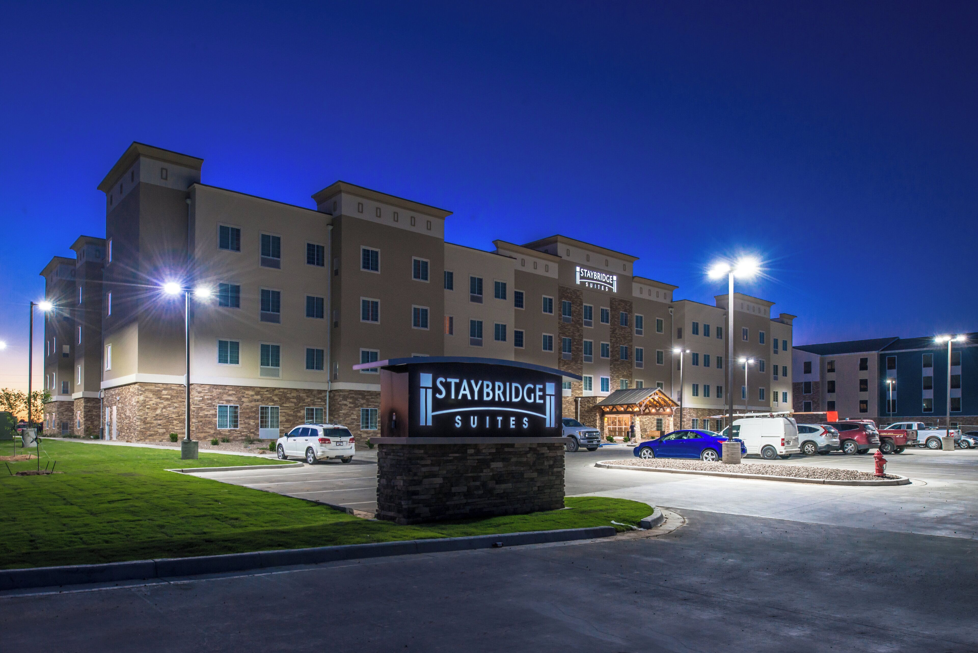 Un hotel Staybridge Suites con un letrero, estacionamiento y varios vehículos.