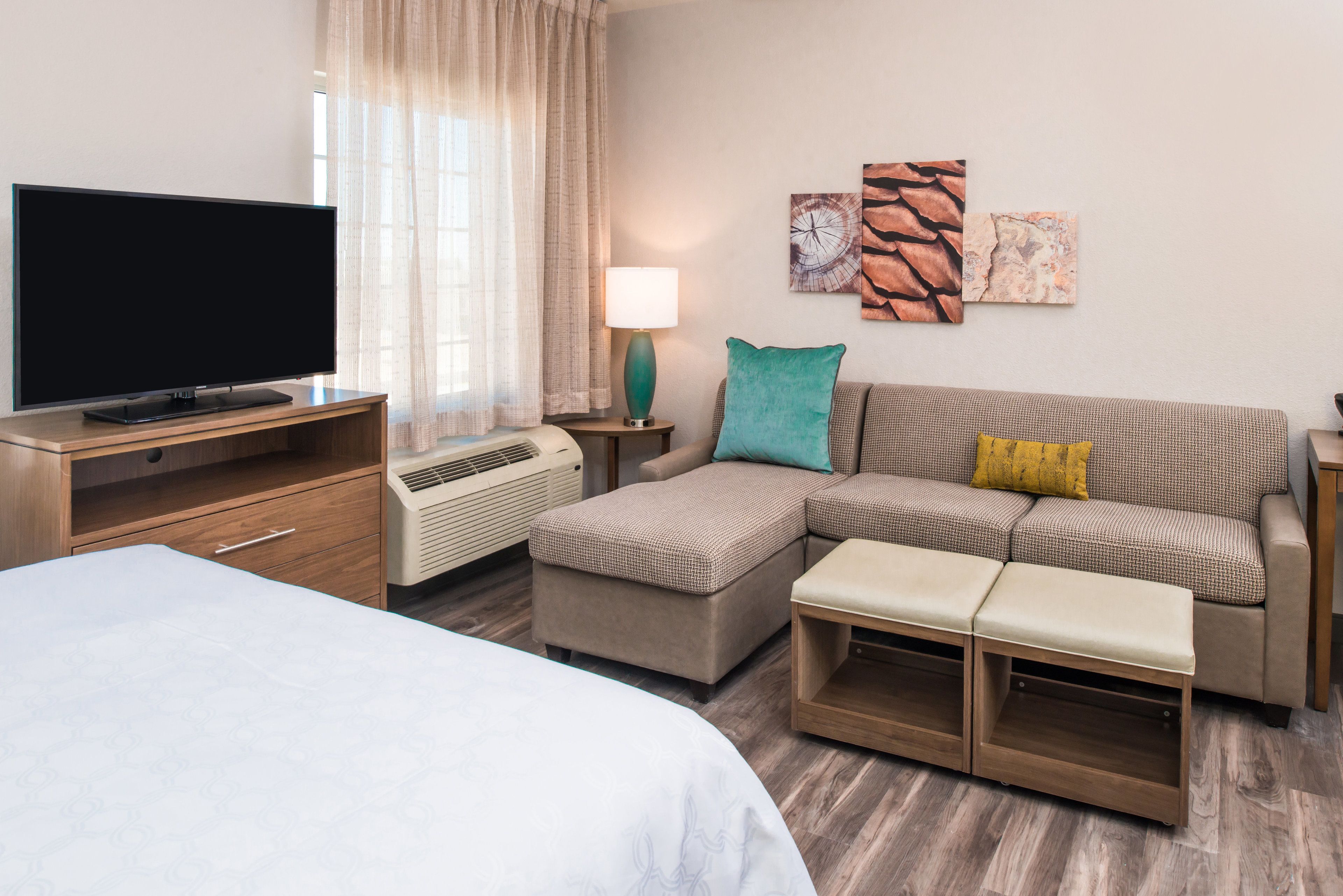 A modern hotel room with a flat-screen TV, a sofa, a bench, a desk, and a lamp.