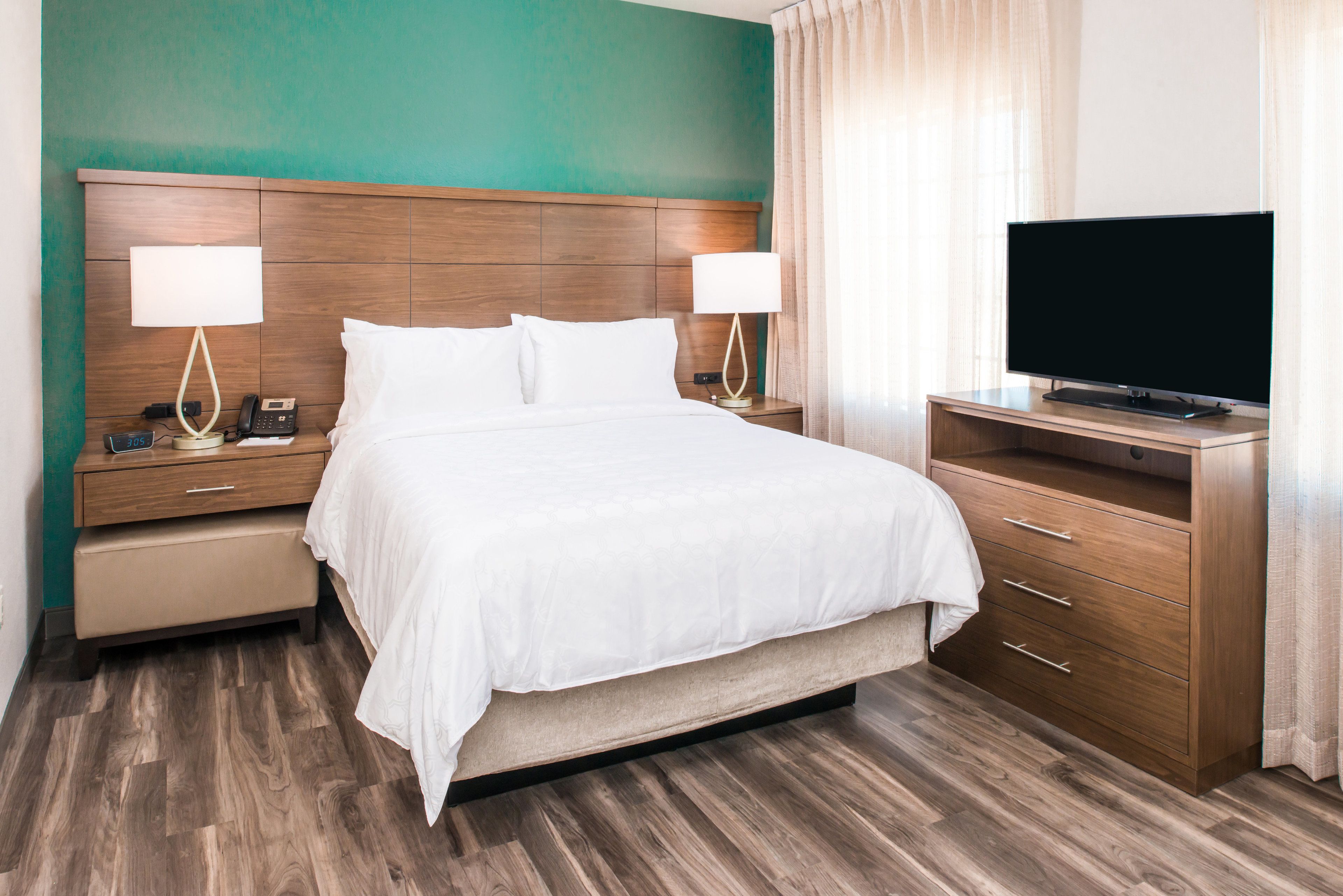 Una habitación de hotel con cabecera de madera, una cama con ropa blanca, dos mesitas de noche con lámparas, un televisor sobre un mueble de madera y piso de madera.