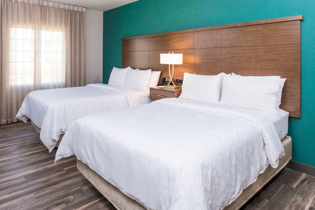 Une chambre d’hôtel avec deux lits, une tête de lit en bois, une table de chevet avec une lampe et une fenêtre avec des rideaux.