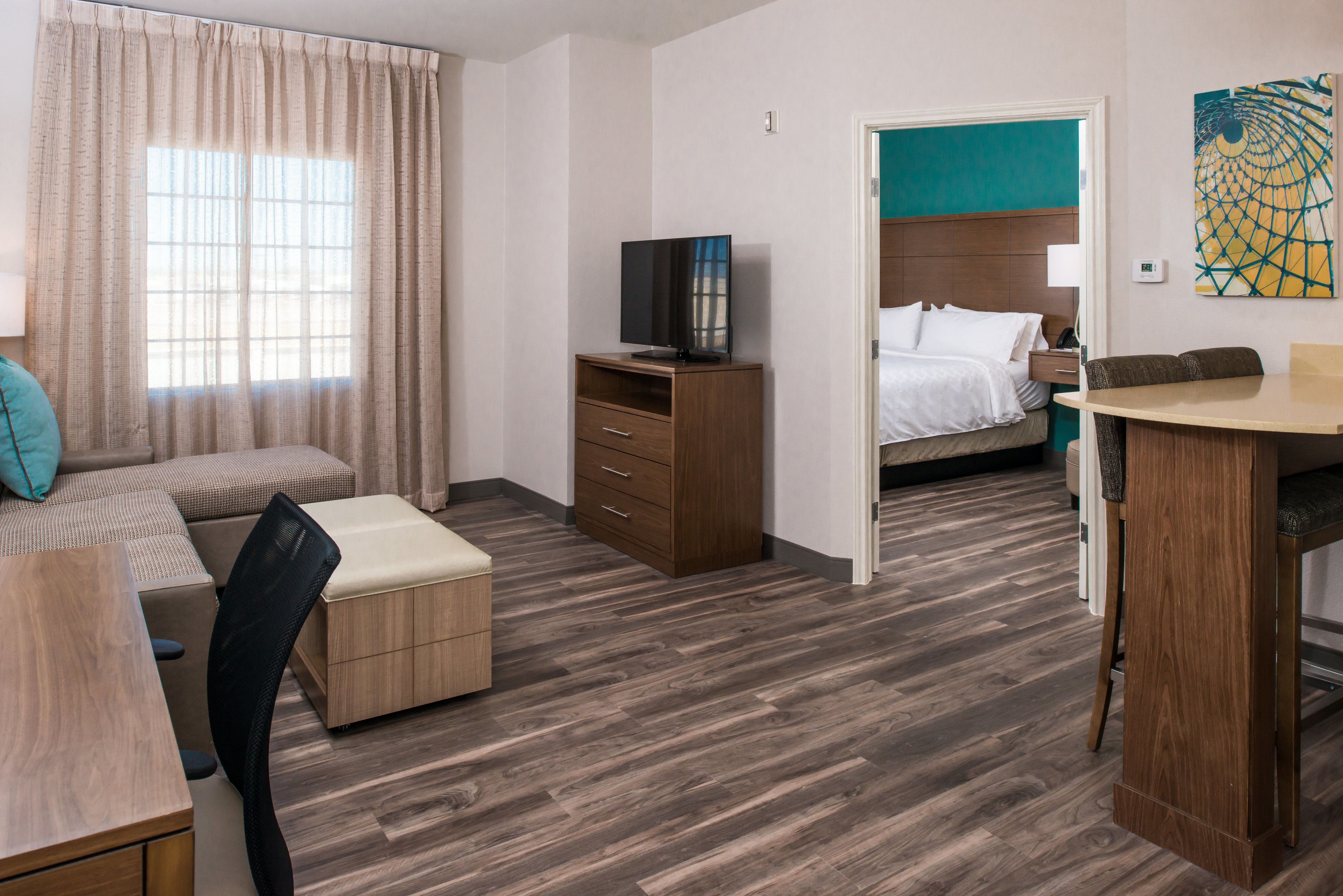 Una habitación de hotel con piso de madera, una cama con ropa blanca, un televisor sobre un mueble de madera, un escritorio con una silla y una ventana con cortinas.