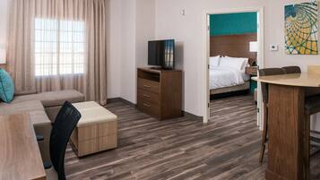 Una habitación de hotel con piso de madera, una cama con ropa blanca, un televisor sobre un mueble de madera, un escritorio con una silla y una ventana con cortinas.