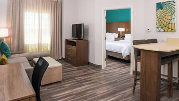 A hotel room with a wooden floor, a TV on a wooden stand, a bed with white linens, a dining table with chairs, and a painting on the wall.