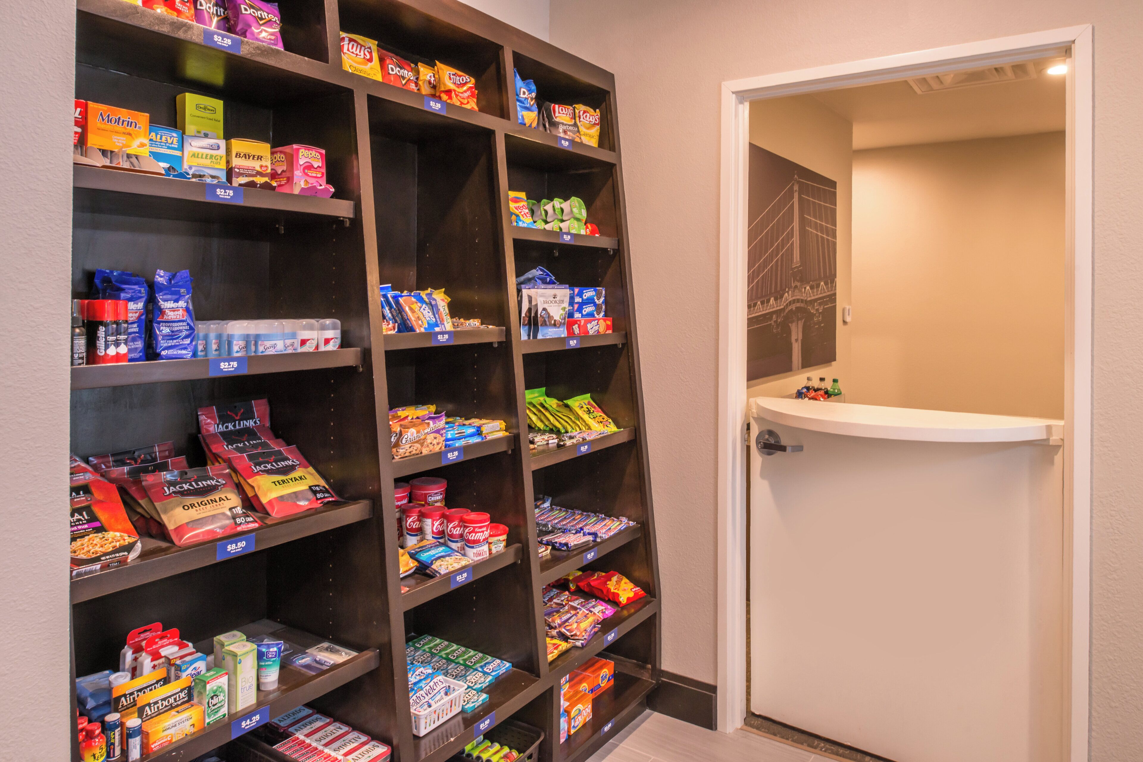 A pantry stocked with snacks and beverages, a door leading to a bathroom, and a framed picture of a bridge on the wall.