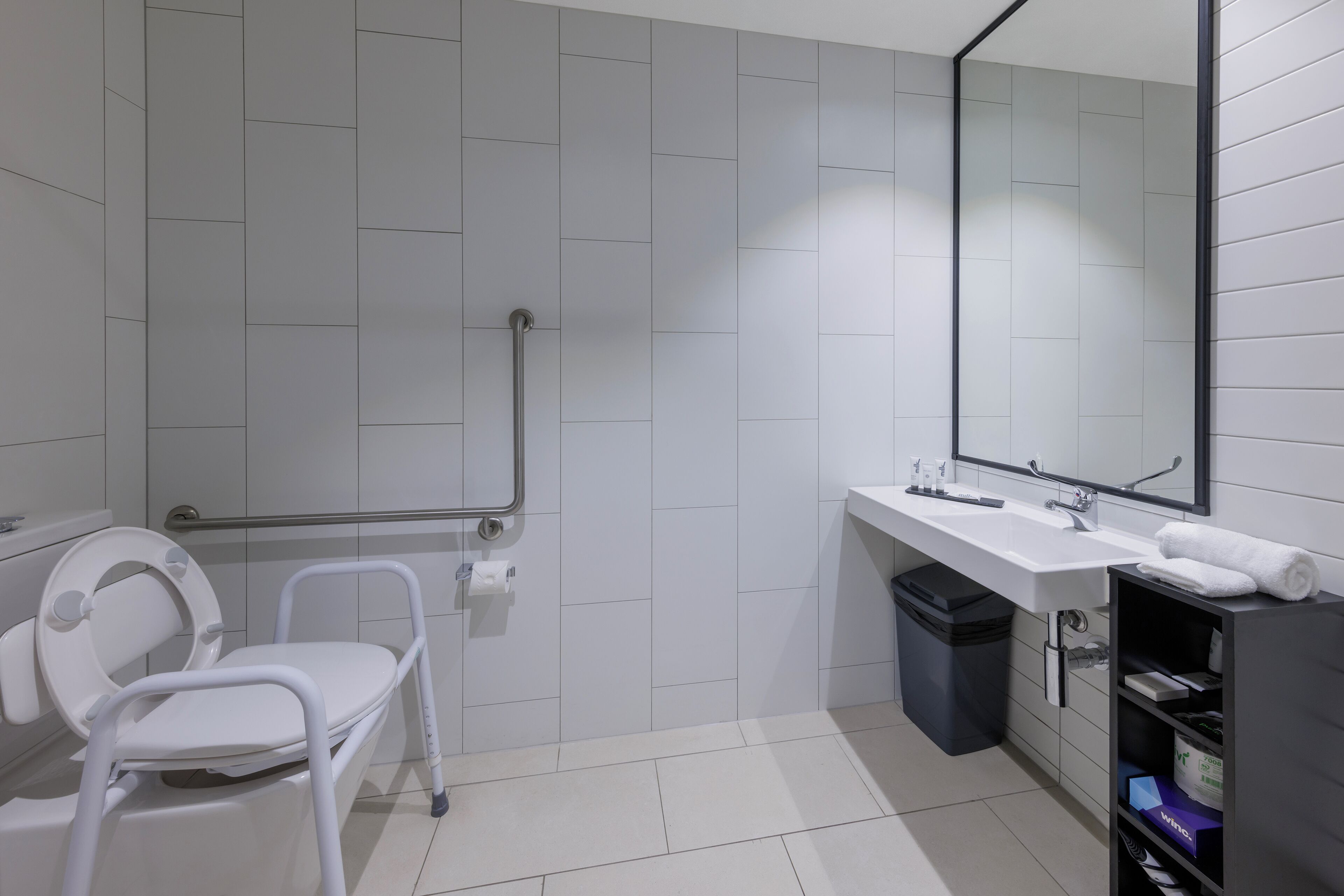 Une salle de bain moderne avec des toilettes blanches, une barre d’appui, un lavabo, un miroir et une poubelle.