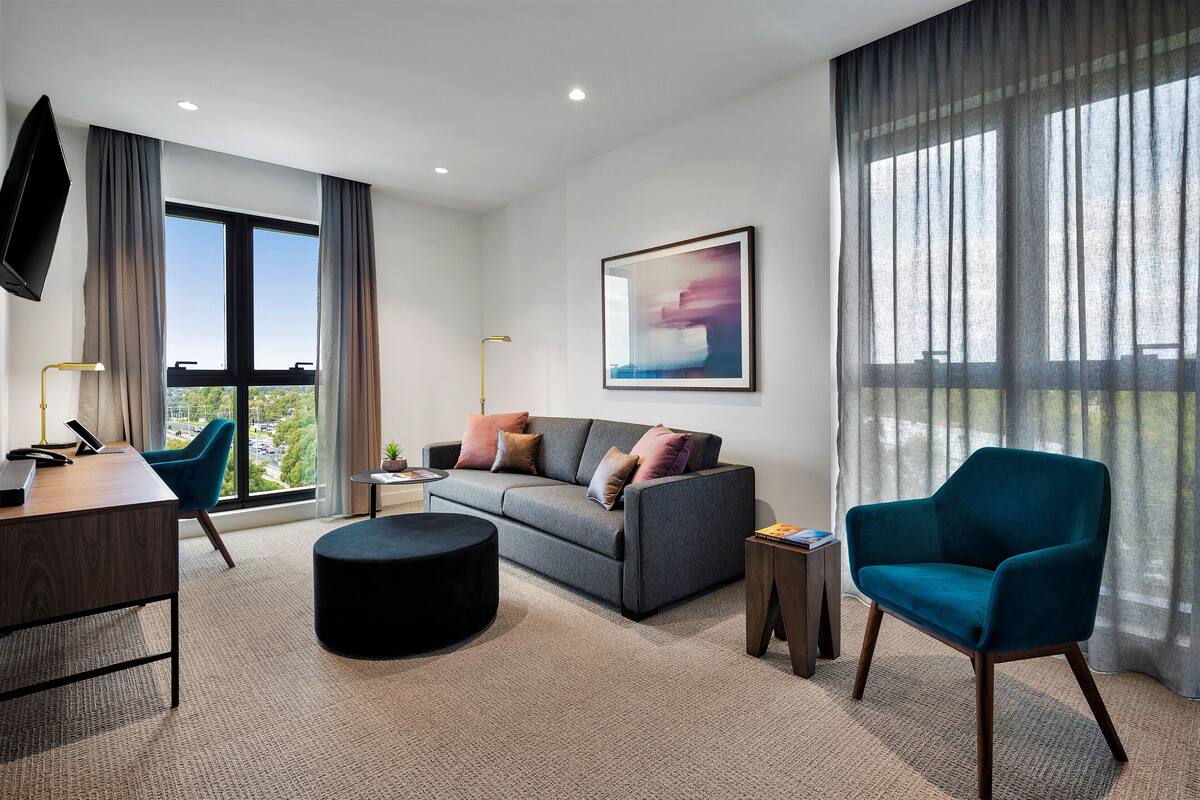 A modern living room with a grey sofa, teal armchair, and a black ottoman. A flat-screen TV is mounted on the wall, and there is a window with curtains.