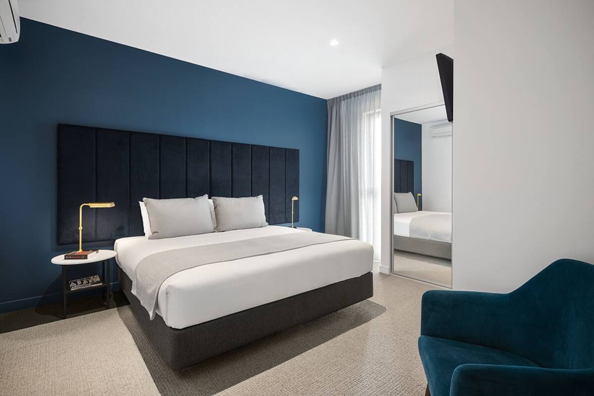 A modern hotel room with a large bed, a nightstand, a desk lamp, a blue armchair, and a large mirror.