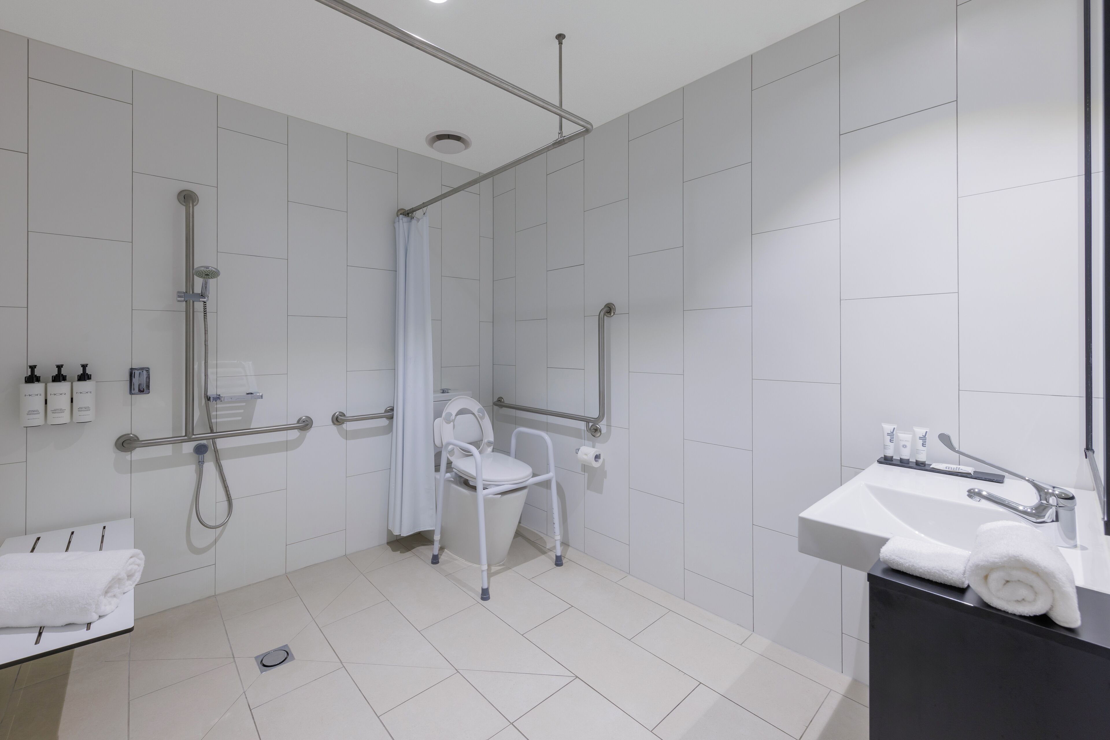 A modern bathroom with a shower, a toilet with a safety seat, a sink with a towel, and a handrail.