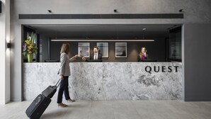 Una reception con la scritta 'QUEST', una persona con una valigia e un/una receptionist.