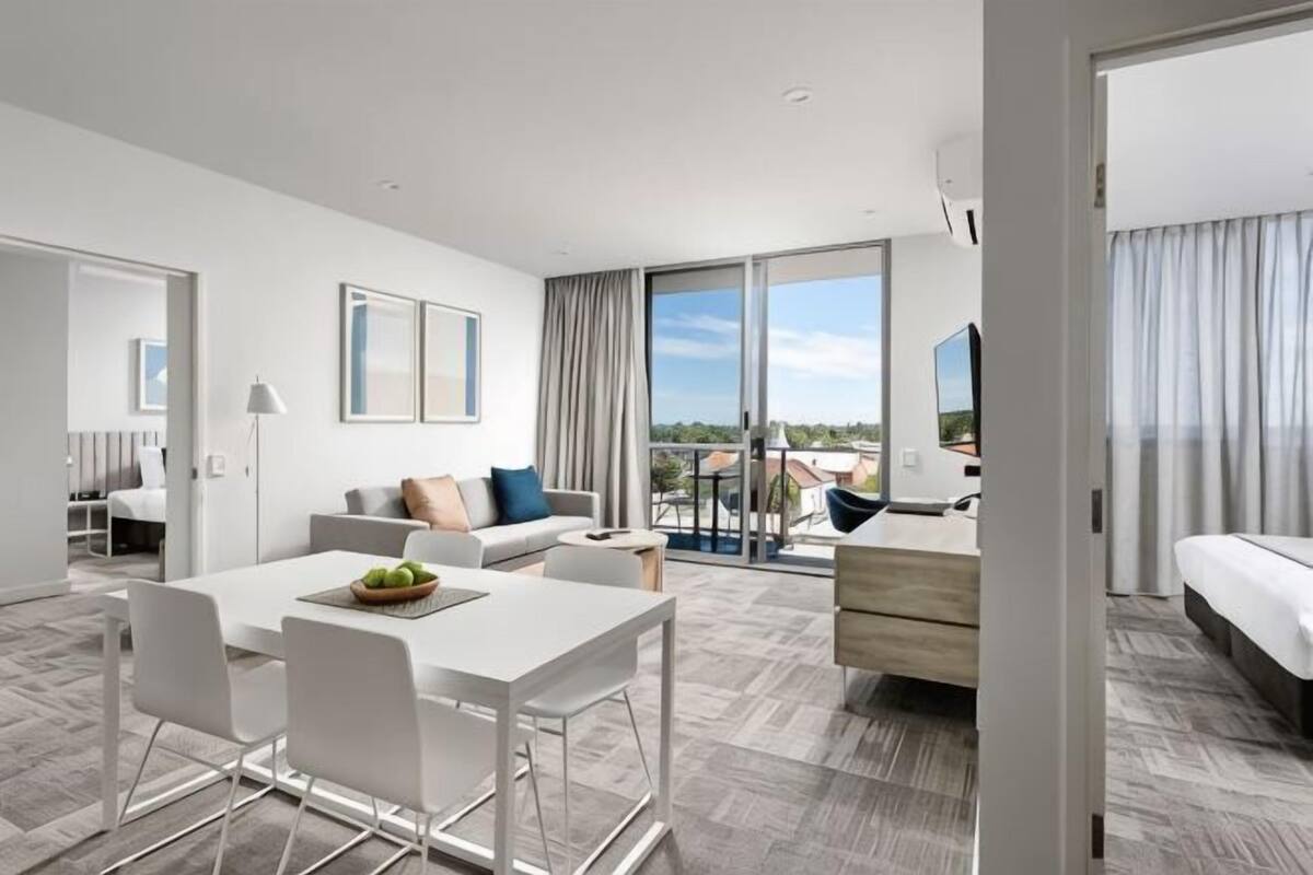 A modern hotel room with a dining area, a sofa, a bed, and a balcony with a city view.
