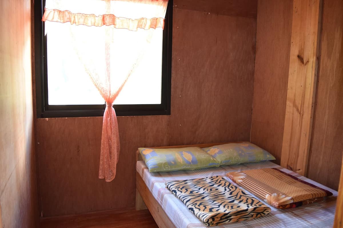 A small wooden room with a single bed, a window with a sheer curtain, and a wooden wall.