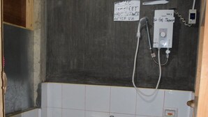 A small, basic bathroom with a toilet, sink, and shower. There is a blue bucket and a sign on the wall.
