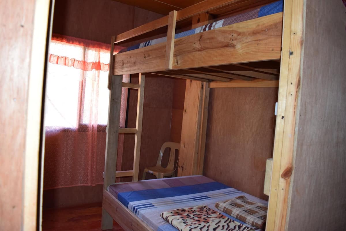 A bunk bed room with a wooden ladder, a single bed with a patterned blanket, and a plastic chair.