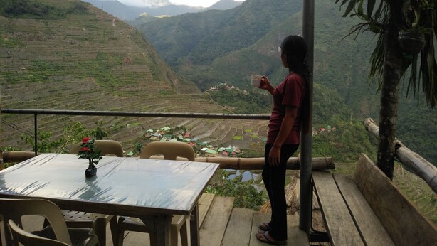A person standing by a table with a view of terraced fields and hills in the background. - Batad View Inn and Restaurant (Banaue)