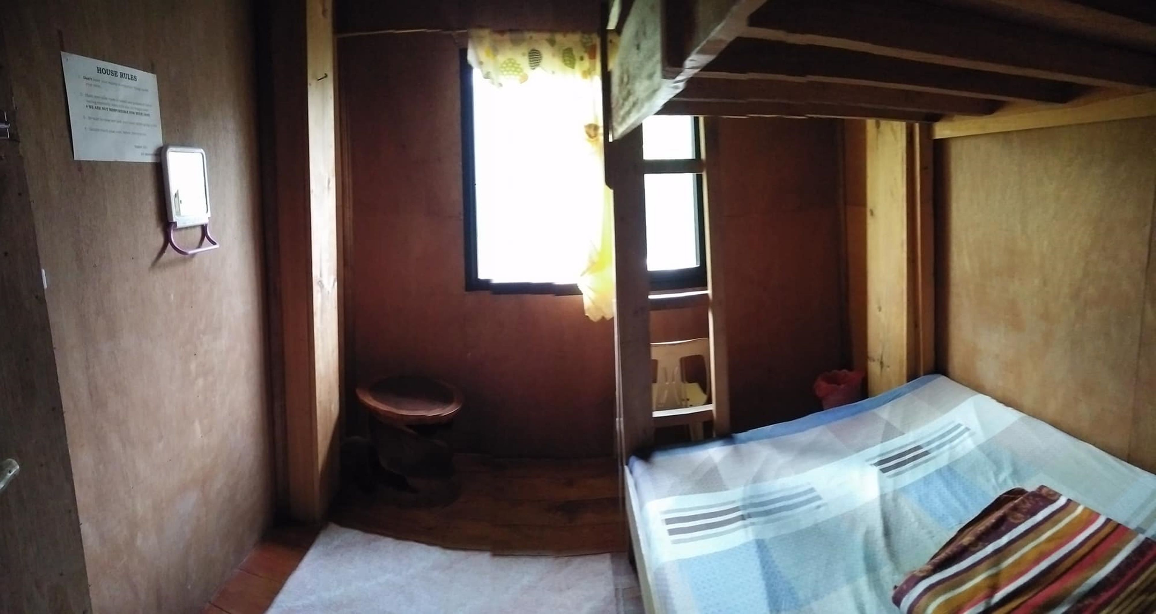 A small, dimly lit room with a single bed, a wooden stool, and a window allowing natural light.