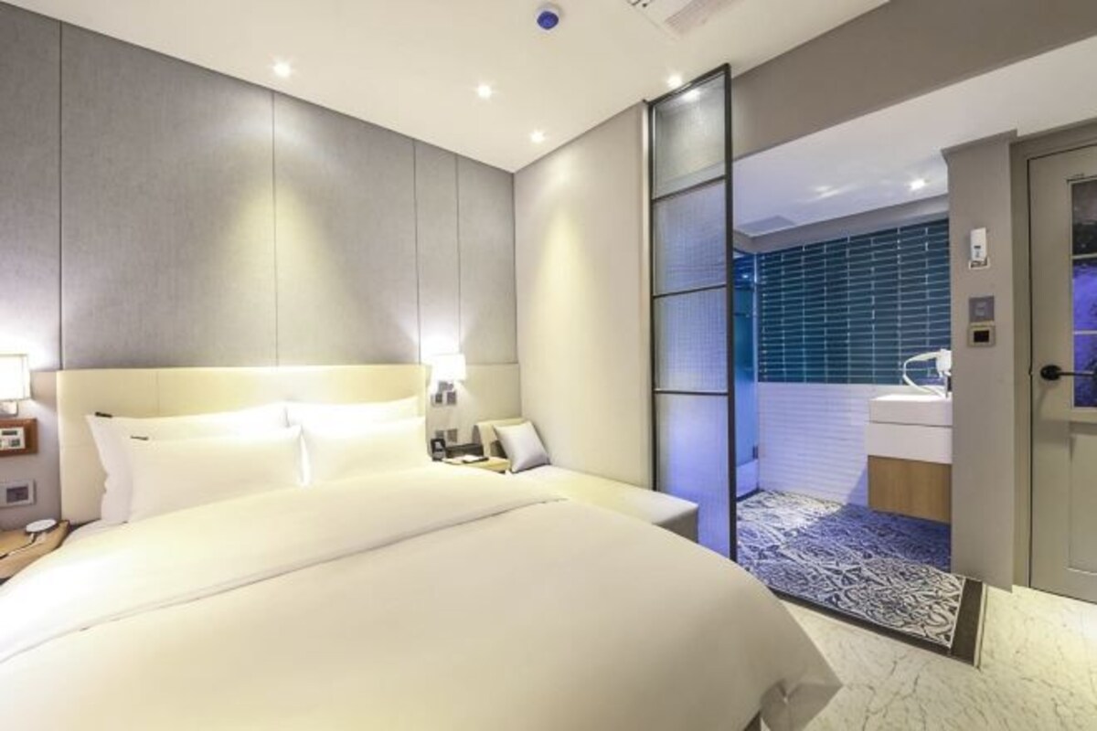 A modern hotel room with a large bed, bedside tables, a nightstand, and a bathroom with a sink and mirror.