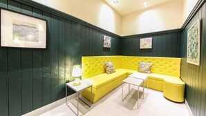 Une pièce d’angle avec un canapé jaune, deux petites tables d’appoint blanches et des murs recouverts de panneaux verts.