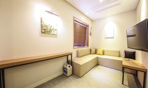 Una sala de estar moderna con un sofá, un televisor, una pequeña mesa, un banco y una ventana con persianas.