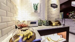 Un espace d’accueil d’hôtel au design moderne, comprenant un mur recouvert de carreaux blancs, une horloge numérique, une machine à café et un panier de fruits.