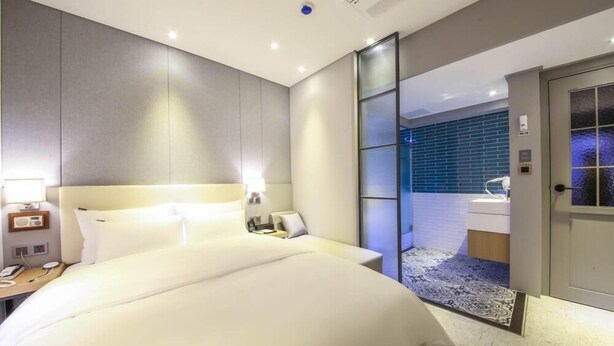 Une chambre d’hôtel moderne dotée d’un grand lit, de tables de chevet, d’une petite table d’appoint, d’une salle de bain séparée par une paroi en verre et d’un petit coin salon.